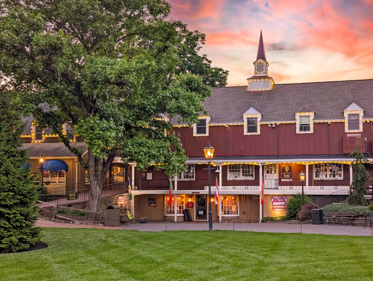 clickbizhub dramatic sunset view of peddler's Village featuring colonial buildings with red siding, church steeple, string lights, and warm window glow against spectacular orange and pink sunset sky with large mature trees silhouetted in foreground