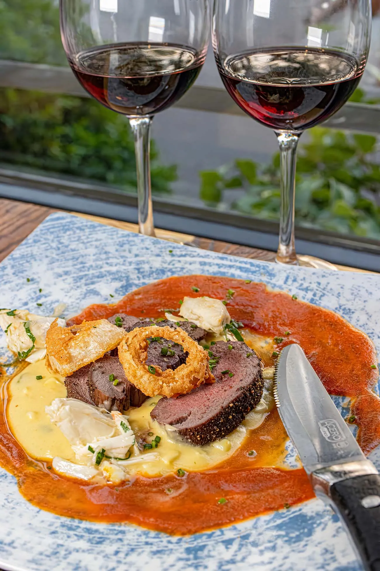 clickbizhub perfectly sliced venison tenderloin with juniper berry crust served over corn and poblano porridge with crispy onion garnish and red wine reduction sauce paired with red wine glasses