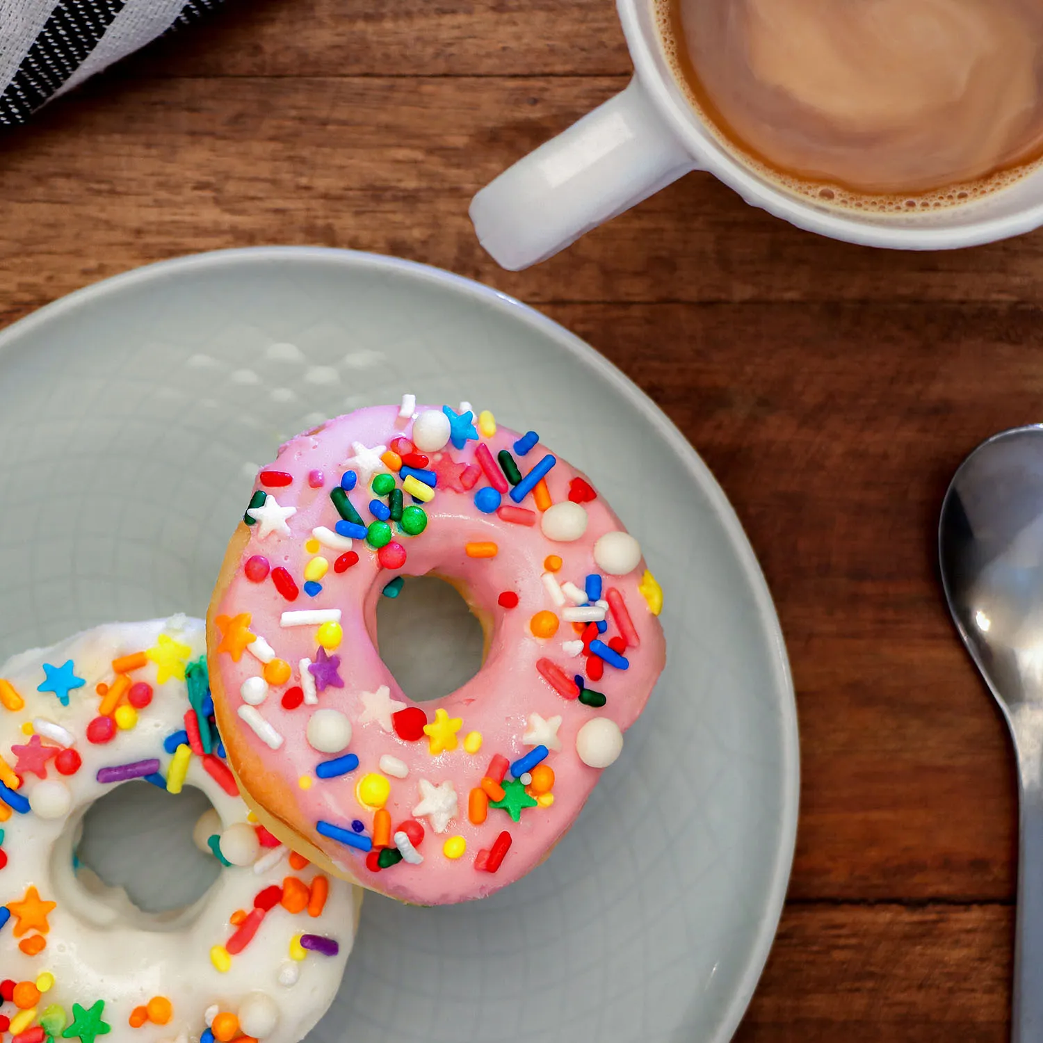 clickbizhub two glazed air fried donuts with rainbow sprinkles on a light blue ceramic plate next to a white coffee mug filled with latte photographed from above on a wooden table surface