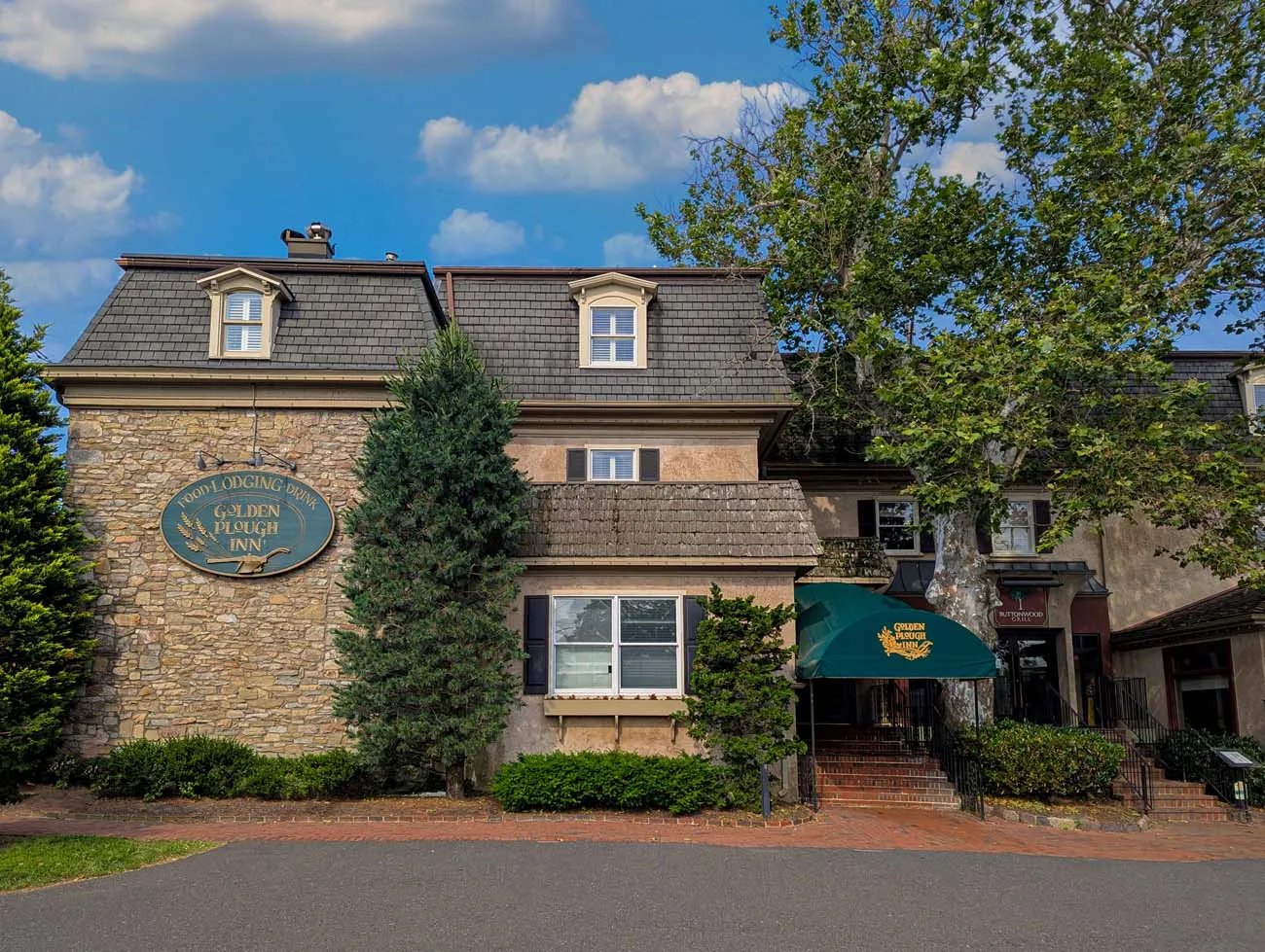 clickbizhub exterior view of the golden plough inn main building featuring colonial architecture with stone and stucco facade green awning dormer windows and surrounded by mature trees and landscaping