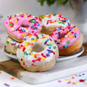 Multiple air fryer donuts with pink and white glazes covered in rainbow sprinkles arranged on a white marble cutting board, with jars of colorful sprinkles visible in the background and plain unglazed donuts to the side.
