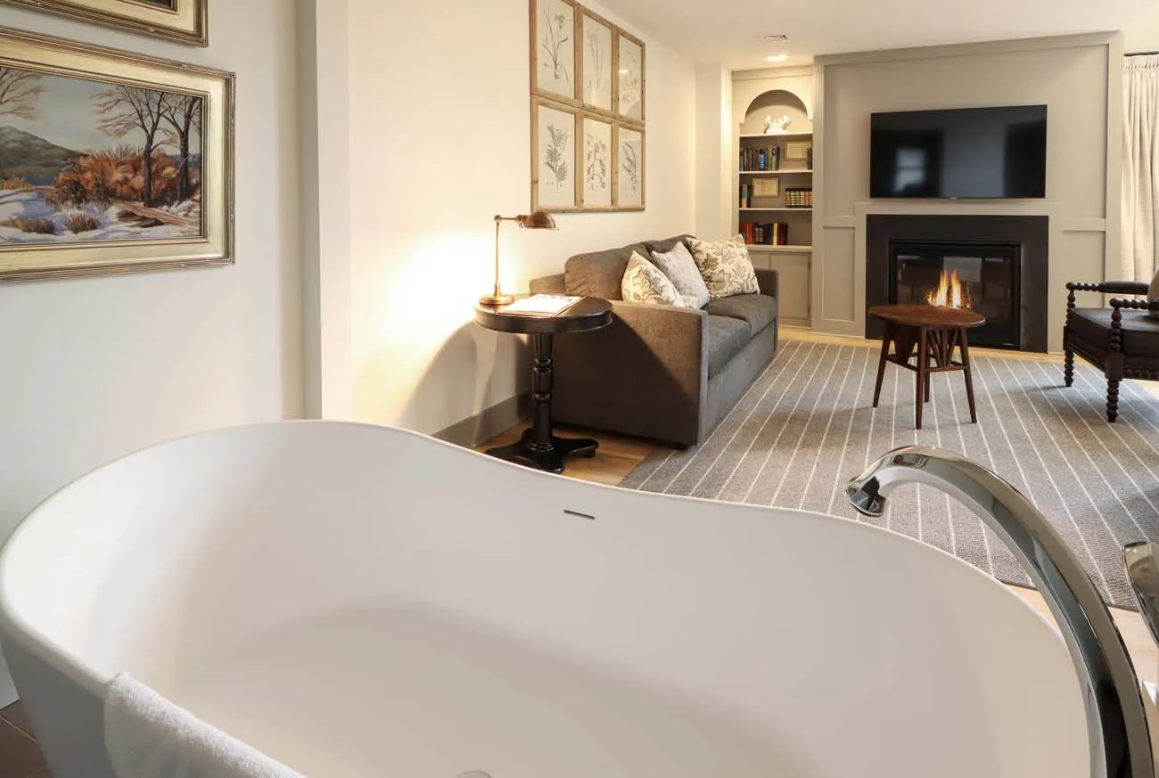 clickbizhub luxurious hotel suite featuring large white soaking tub in foreground comfortable seating area with gray sofa gas fireplace built in bookcases flat screen tv and botanical artwork on cream colored walls