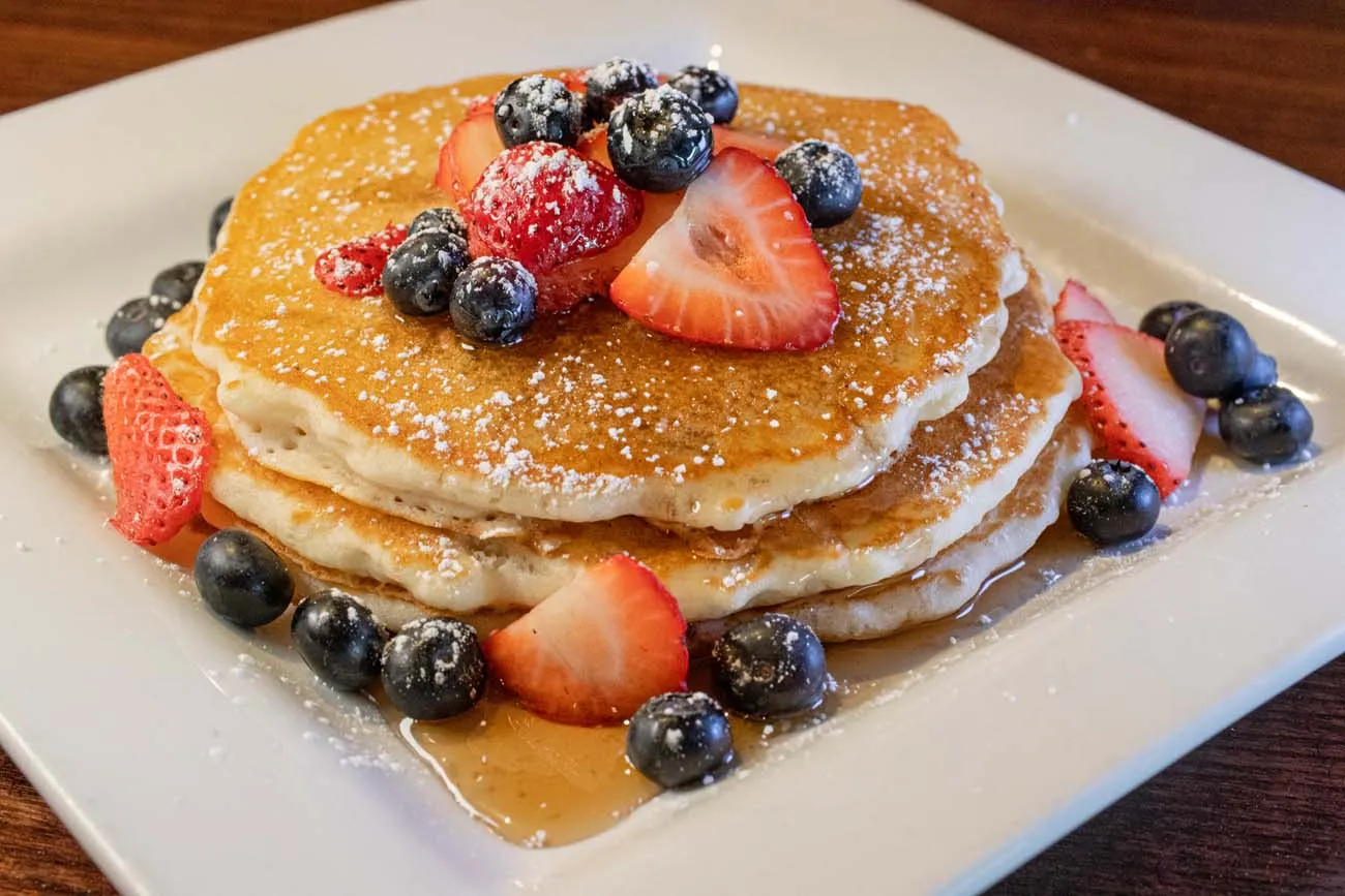clickbizhub stack of golden pancakes topped with fresh strawberries blueberries raspberries powdered sugar and maple syrup being poured from above served on a white plate at buttonwood grill