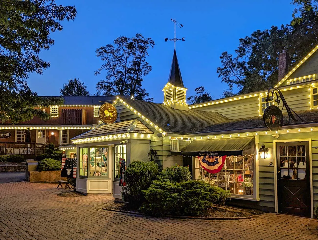 clickbizhub evening view of illuminated colonial style shops at peddler's Village with string lights outlining building edges, Golden Plough Inn sign visible, American flag, and warm glow from shop windows against deep blue twilight sky