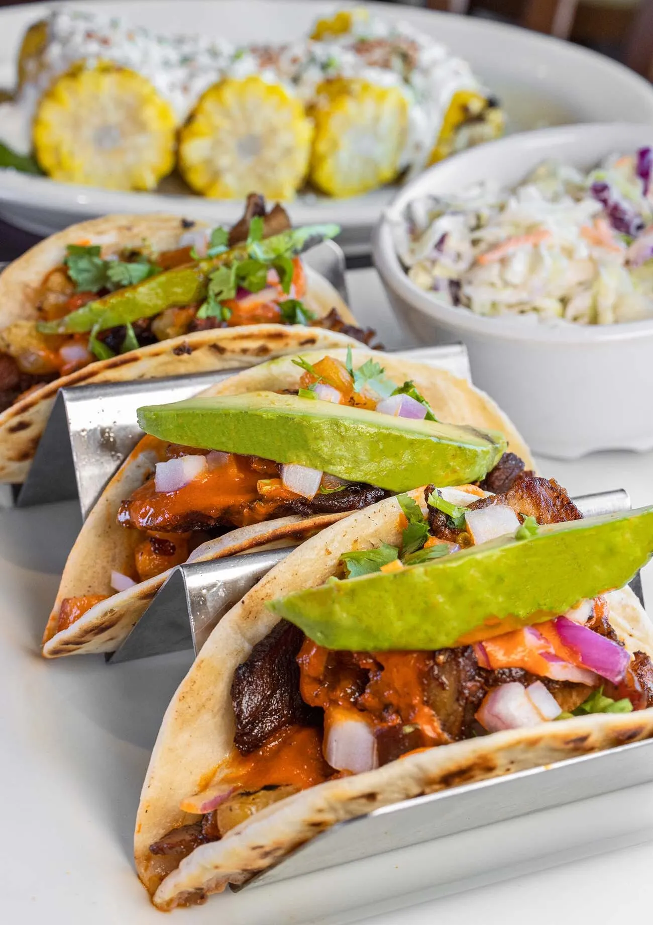 clickbizhub three soft flour tortillas filled with al pastor pork topped with fresh avocado slices diced red onions cilantro and orange sauce served in a metal taco holder alongside grilled corn on the cob and coleslaw at buttonwood grill in peddler's Village