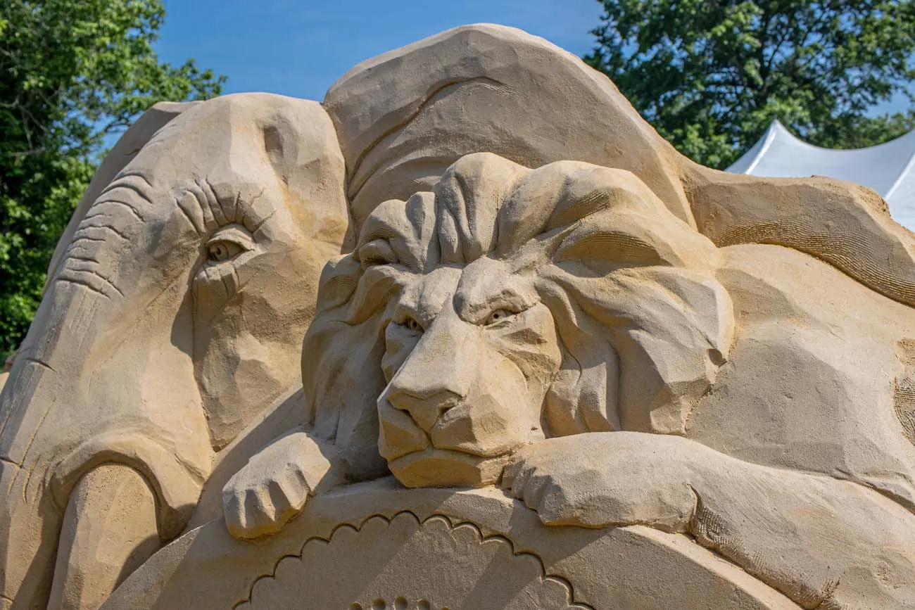 clickbizhub close up view of masterfully carved sand sculpture showing a majestic lion's head and mane alongside an elephant, displaying intricate texture work and realistic facial features against clear blue sky