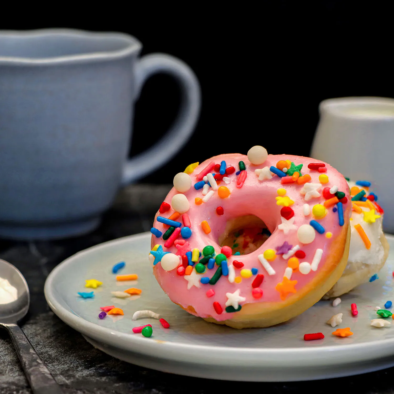 clickbizhub single pink glazed air fryer donut with multicolored sprinkles on a white plate with scattered sprinkles around it alongside a blue coffee mug in soft focus background showing the donut's air fried fluffy texture.