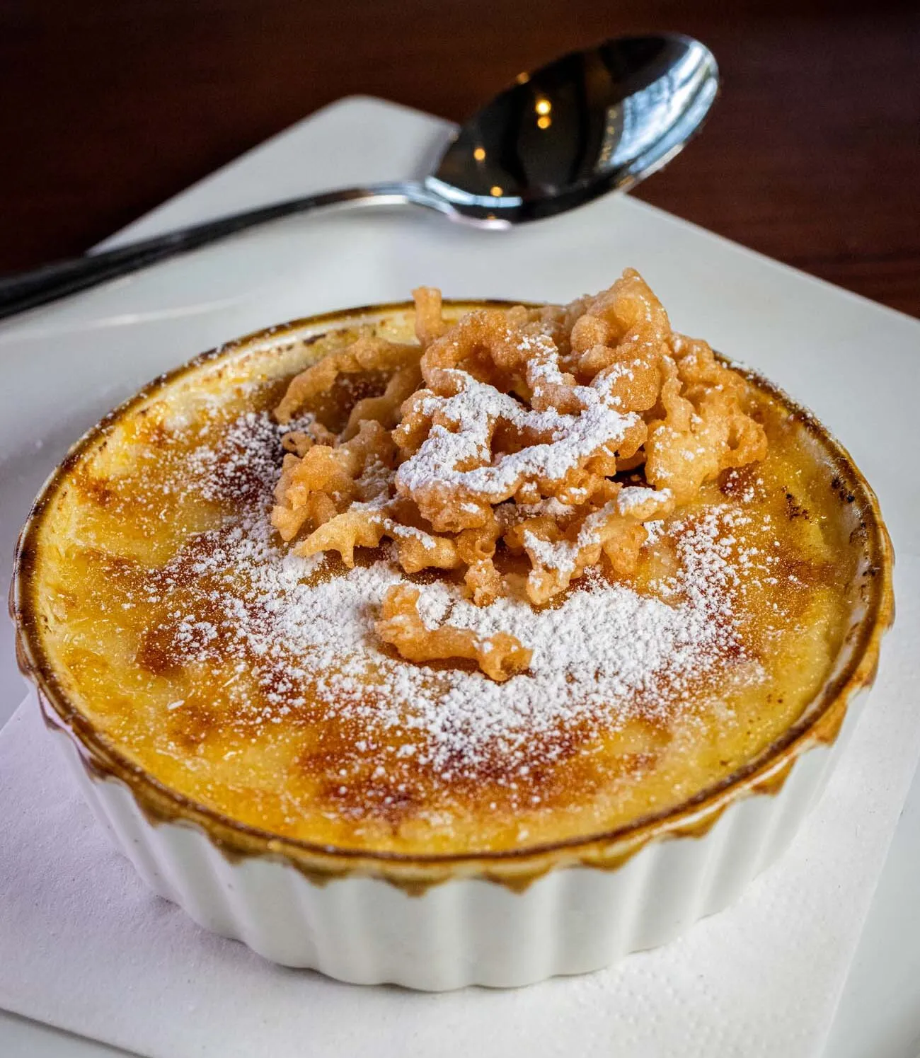 clickbizhub layered funnel cake crème brûlée dessert served in a glass with whipped cream crumble pieces and caramelized sugar cubes on top being eaten with a spoon at cock n bull